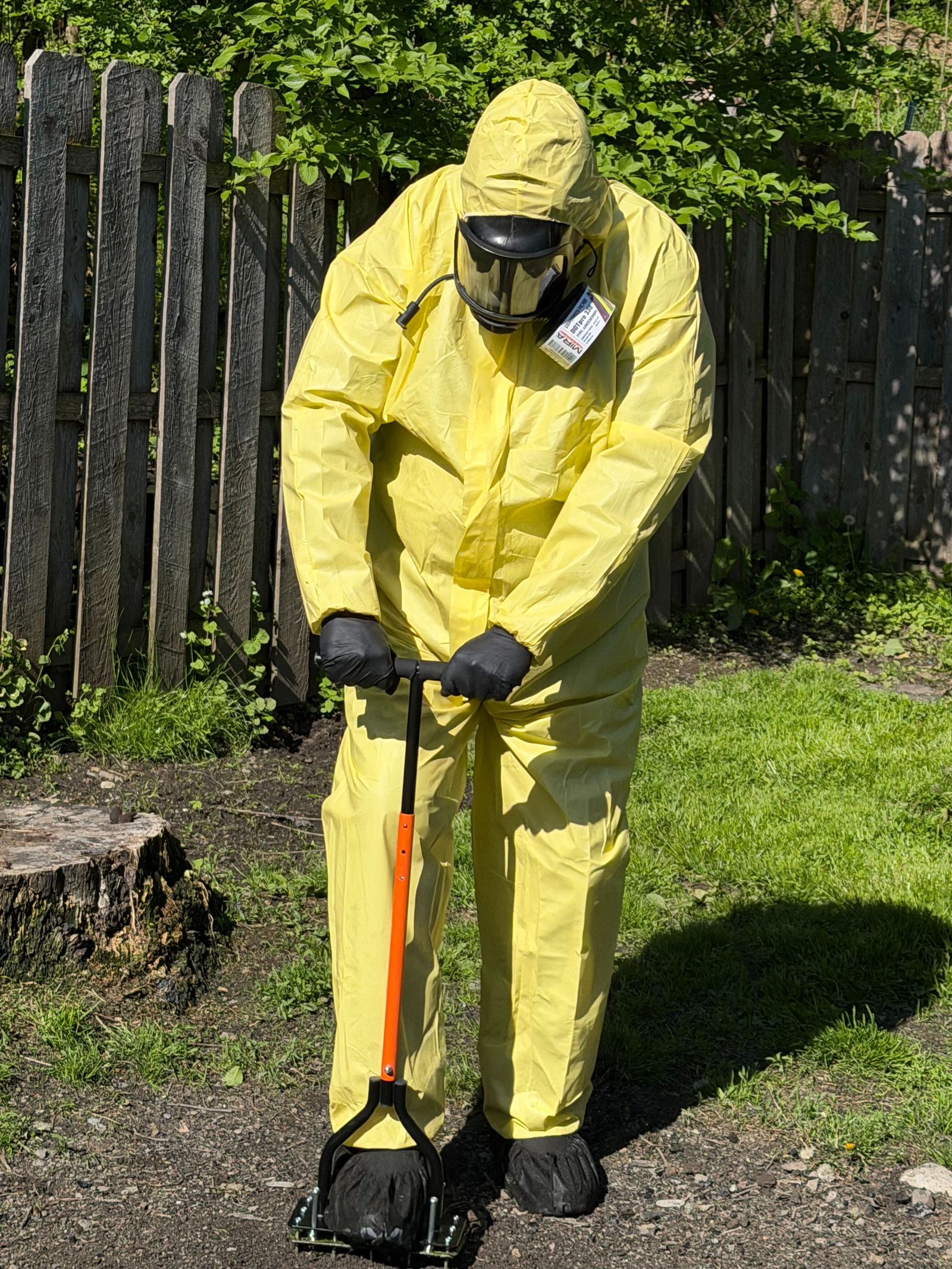An applicator in a protective suit and respirator presses a hand aerator into bare soil in a fenced yard.