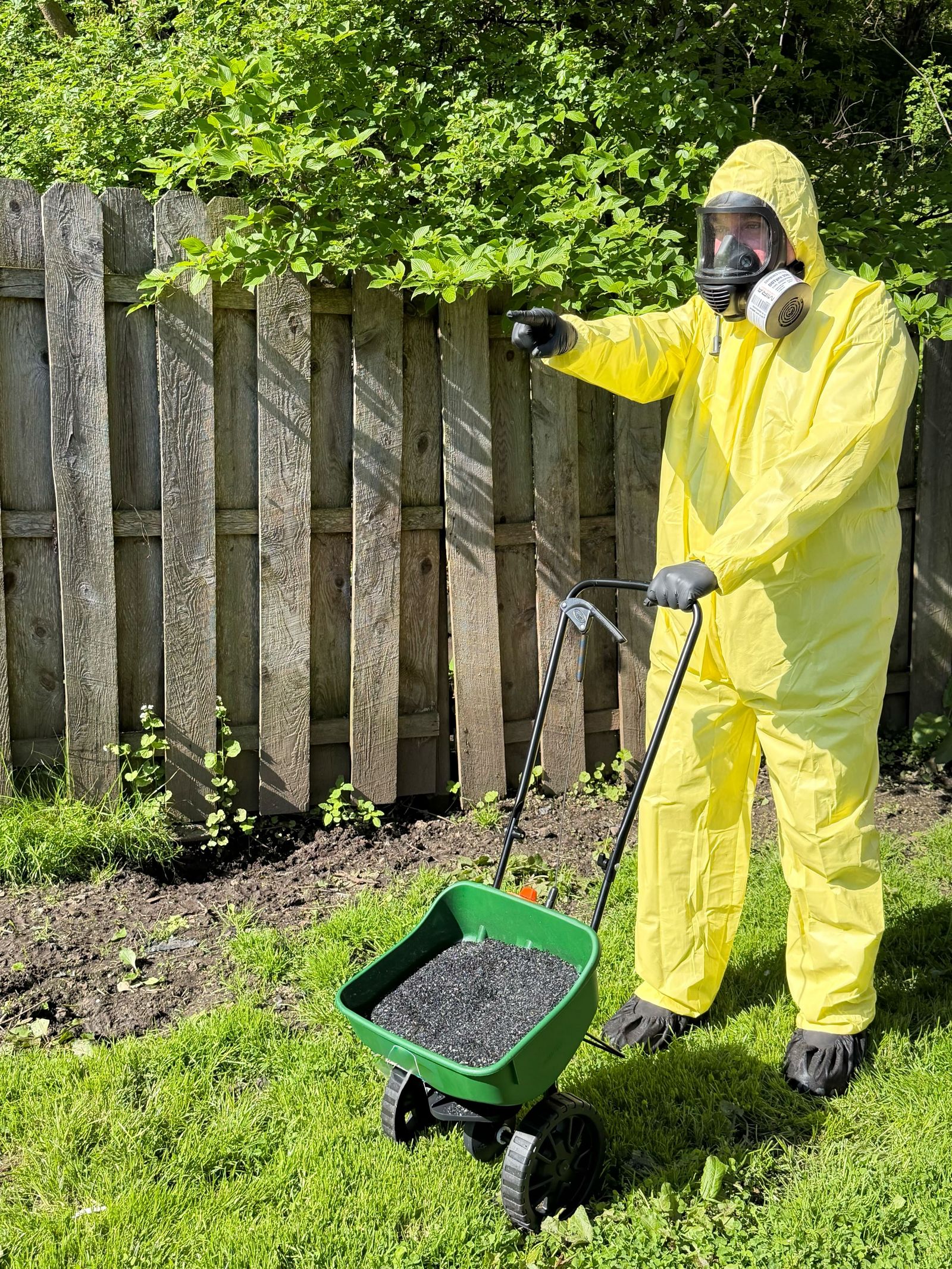 An applicator in a protective suit and respirator beside a broadcast spreader filled with dark granular amendment.