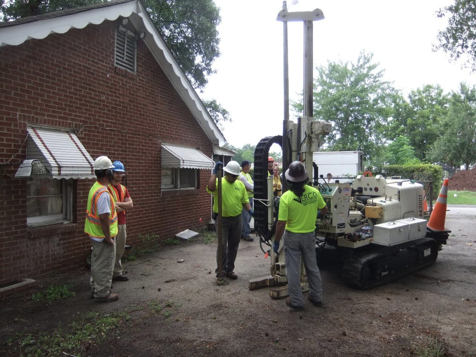 Field crew in safety vests operating a drill rig to collect a soil sample beside a brick building.