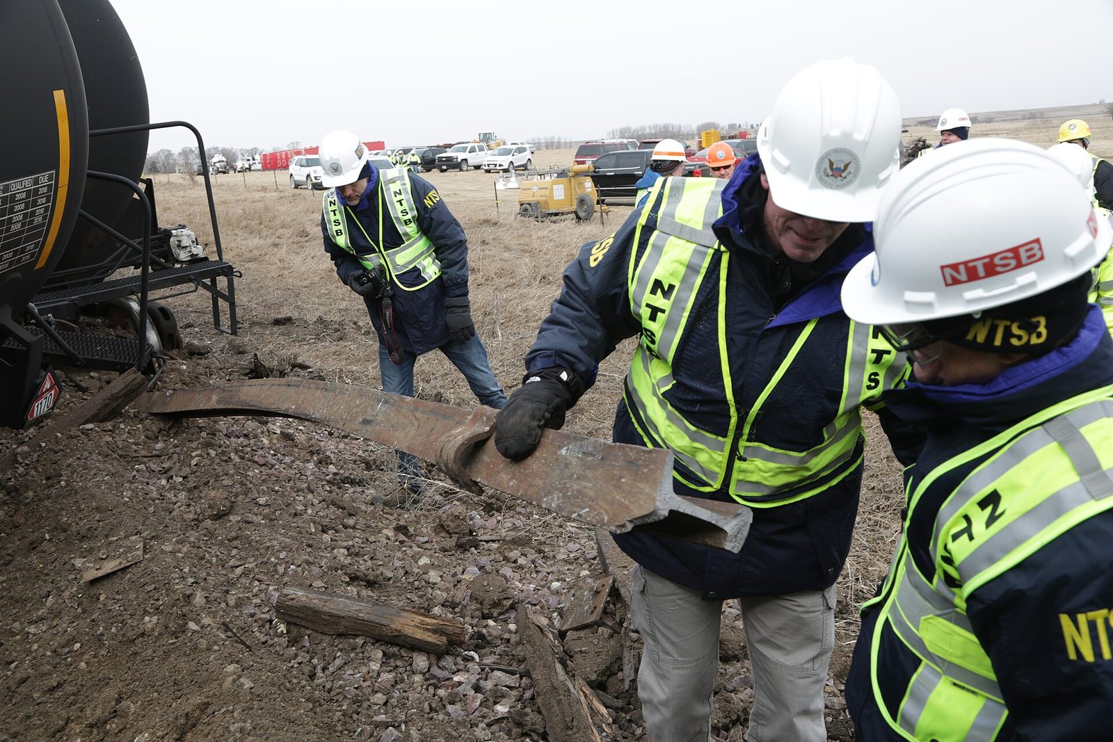 Investigators in hard hats and safety vests examine ground beside a derailed rail car.